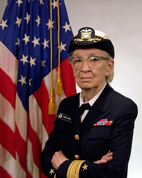 Portrait of Grace Hopper in uniform in front of
                        an USA flag